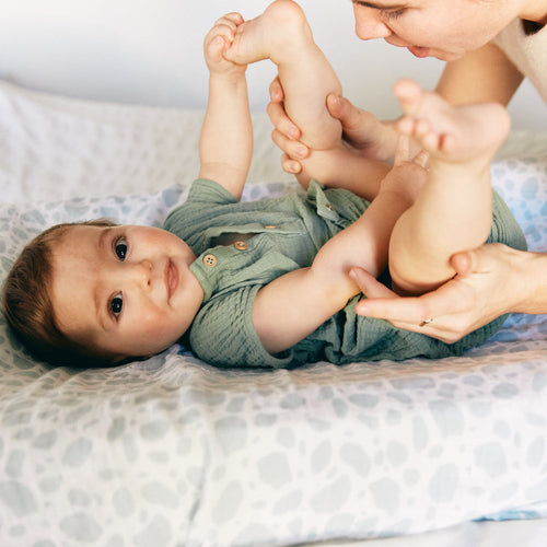 Una bebé feliz estirada en la cama mientras su mamá le ayuda a hacer ejercicios para el estreñimiento - Mustela España - 1