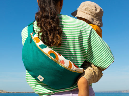 Una madre cogiendo a su hijo frente al mar llevando la riñonera en tonos verdes con estampado de aguacates y flores de Mustela. - Mustela España - 2