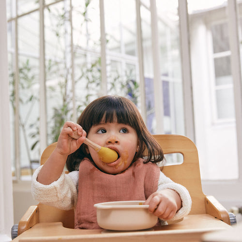 Bebé niña sobre su silla comedor comiendo un pure con una cuchara - Mustela España - 1