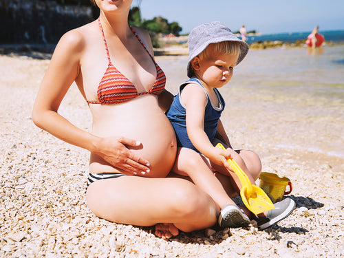 Una madre embaraza con su bebé disfruta de la playa mientras se aplica correctamente el protector solar de Mustela  - Mustela España - 1