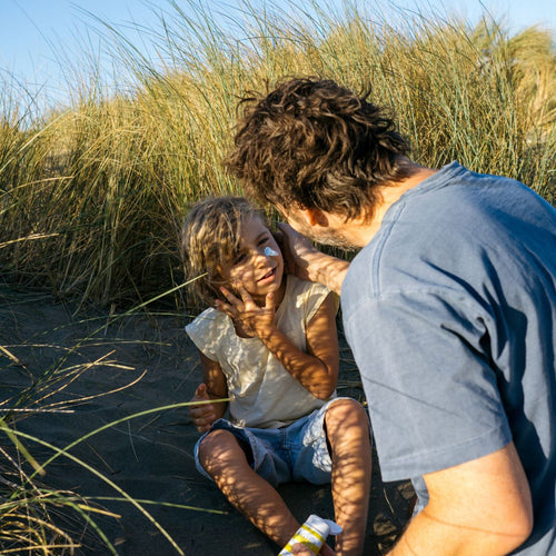 Padre junto su hijo que se esta aplicando crema solar en la cara entre dunas y hierbas altas en un día soleado. - Mustela España - 1