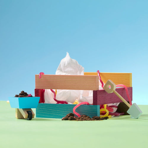 Escena colorida en miniatura con una caja de madera pintada, una servilleta blanca sobresaliendo, tiras decorativas de colores, una carretilla azul con tierra y pequeñas herramientas de jardinería sobre un fondo azul y verde. - Mustela España - 1