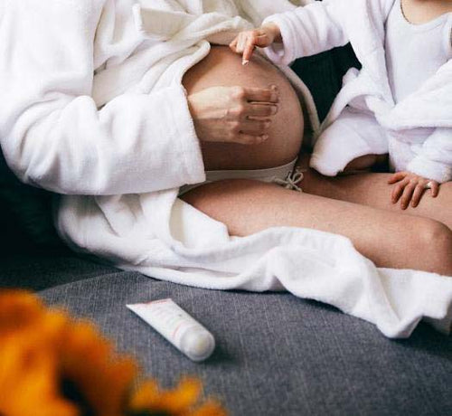 Una mamá embarazada y su hija, las dos con albornoz, después de tomar un baño y aplicándose crema Mustela en la barriga - Mustela España - 1