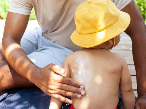 un padre aplica protector solar en la espalda de su niño que lleva un gorro. slider - Mustela España - 3