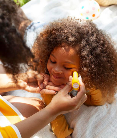 Una madre aplica el stick solar SPF50 Mustela en las mejillas de su niña. 