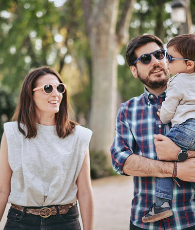 Una familia joven, compuesta por una madre, padre y un bebé, disfruta de un paseo por un parque, llevan gafas Mustela by Parafina en diferentes modelos..